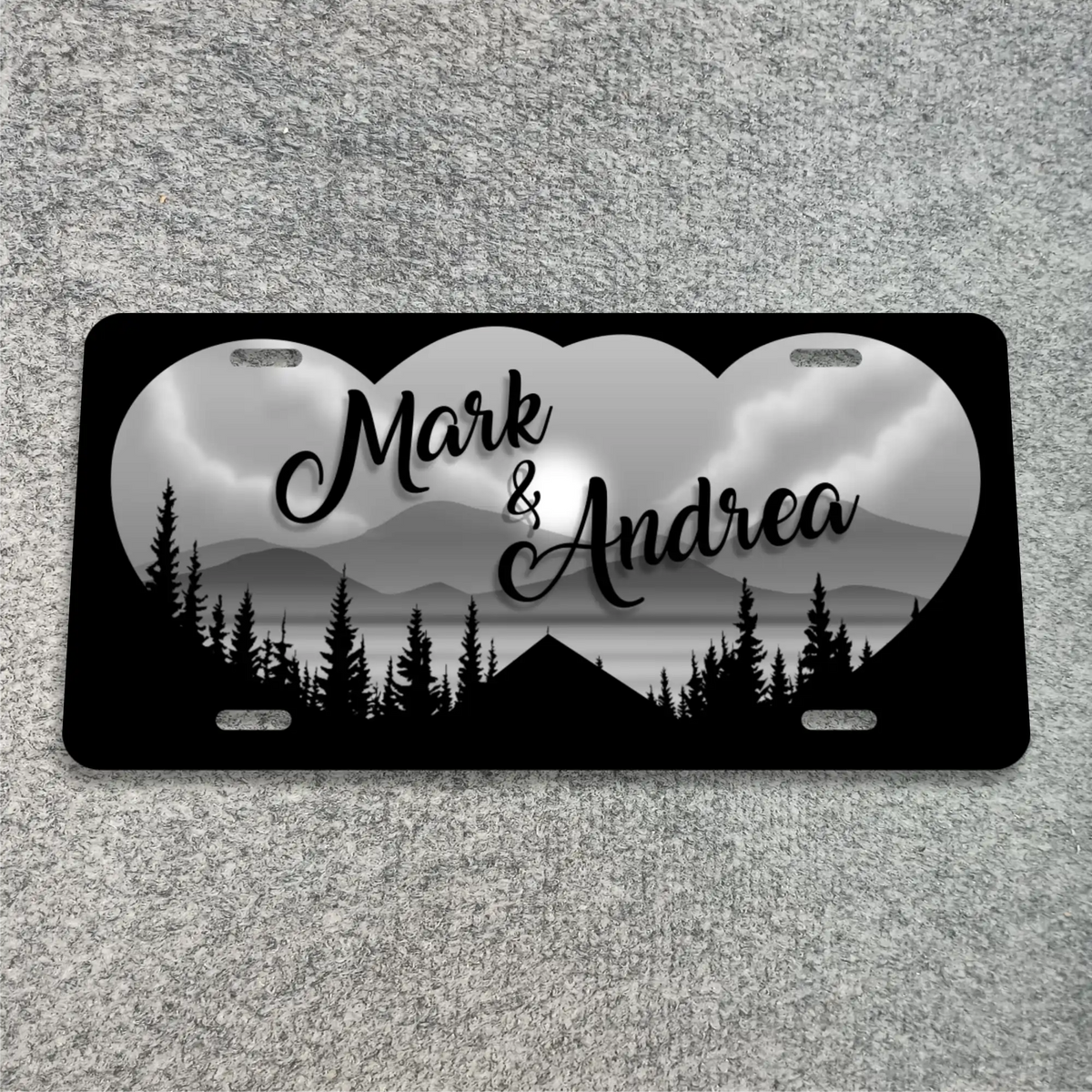 A black license plate with rounded corners, featuring a grayscale heart-shaped mountain and lake landscape with black tree silhouettes. The names 'Mark & Andrea' are printed in black script across the center.