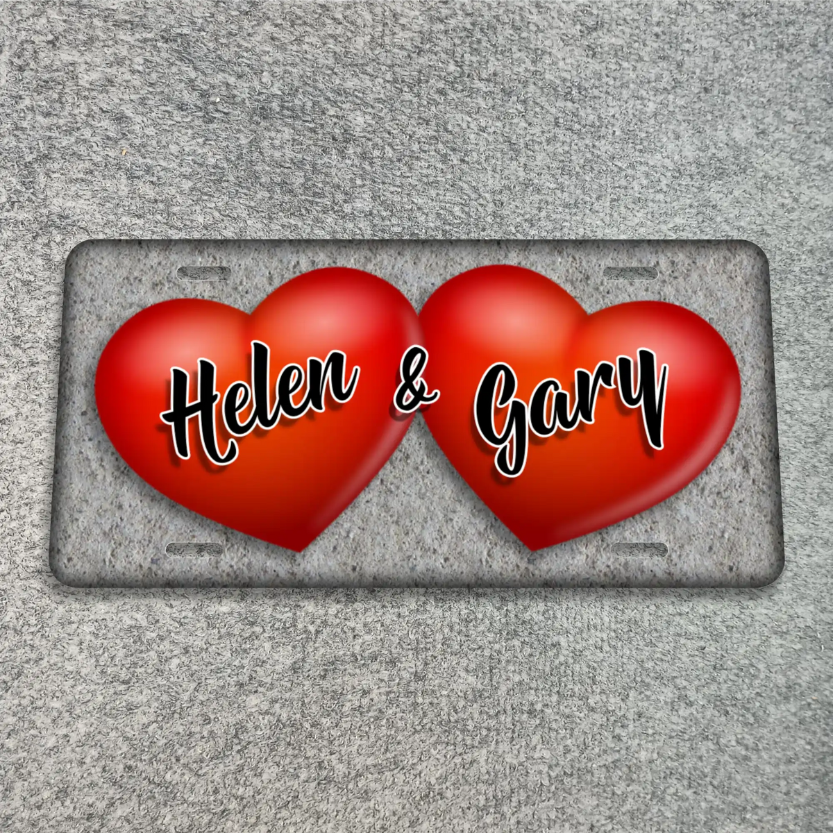 A textured gray license plate featuring two glossy red hearts side by side, with the names 'Helen & Gary' written across the hearts in white cursive with black outlines. The plate is shown on a textured gray background.