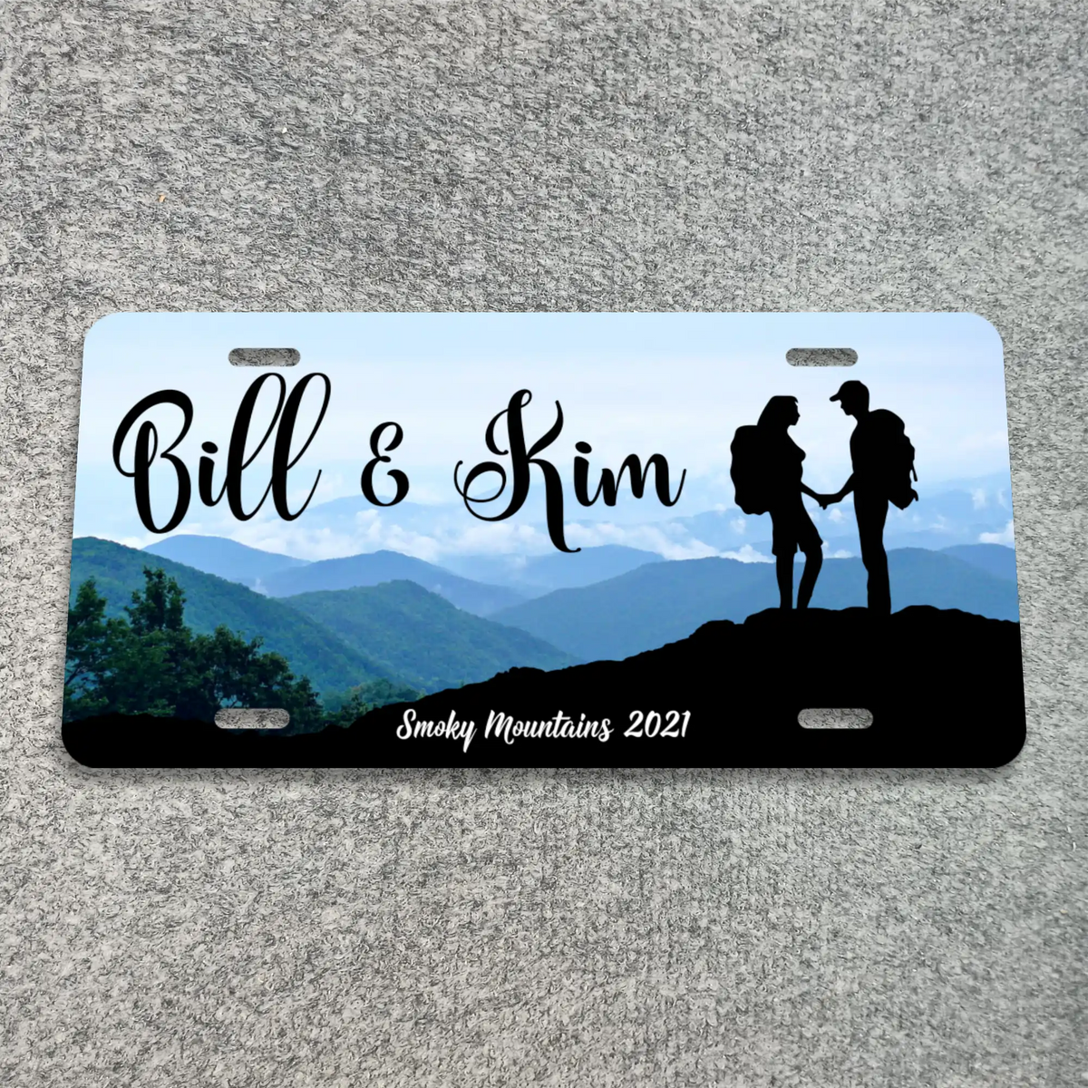 A custom novelty license plate featuring a scenic mountain landscape with the text 'Bill & Kim' and 'Smoky Mountains 2021', plus a black silhouette of a couple holding hands on a hill. The plate is photographed flat on a gray textured surface.