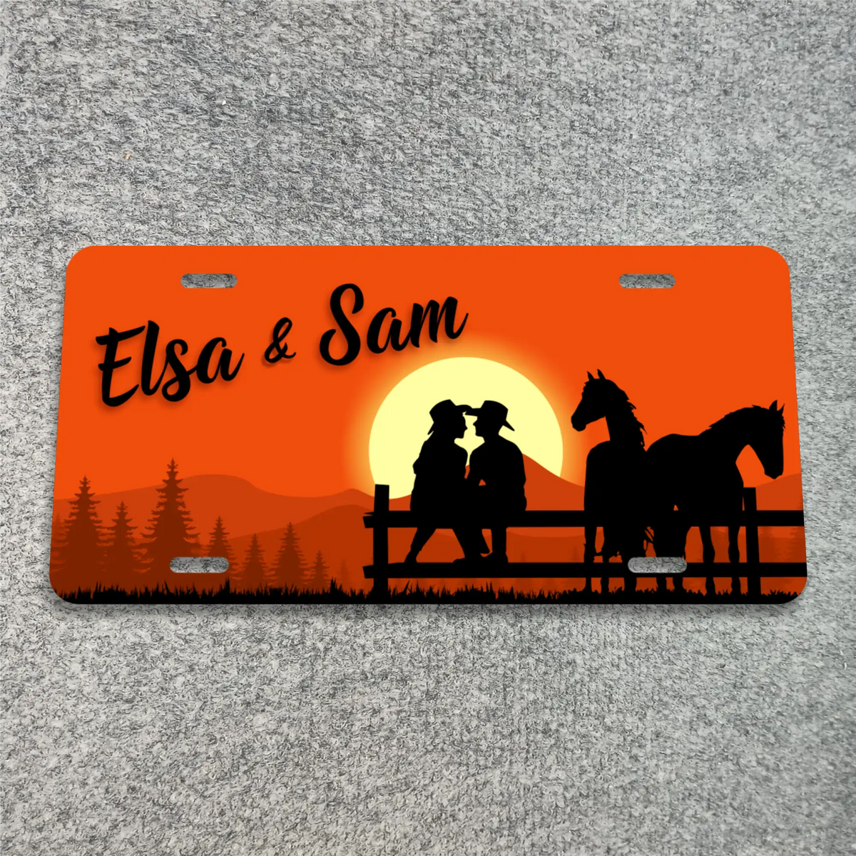 A license plate featuring a sunset design with the text 'Elsa & Sam' in black script, silhouettes of a couple, a horse, trees, and mountains on an orange background, displayed on a gray textured surface.