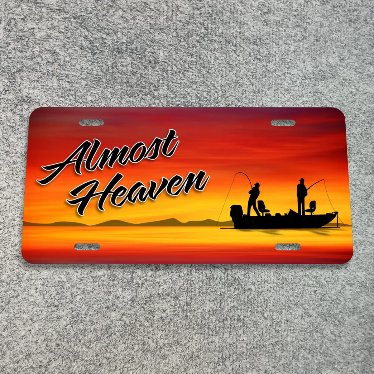 A license plate with rounded corners, featuring a sunset fishing scene and the text 'Almost Heaven' in black script. The plate is photographed flat on a gray textured surface.