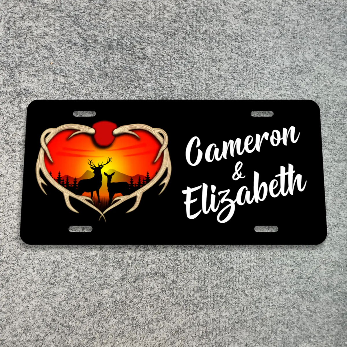 A black license plate featuring a heart-shaped antler border with a deer silhouette against a red and orange sunset, alongside the personalized names 'Cameron & Elizabeth' in white script. The plate is photographed flat on a textured gray background.