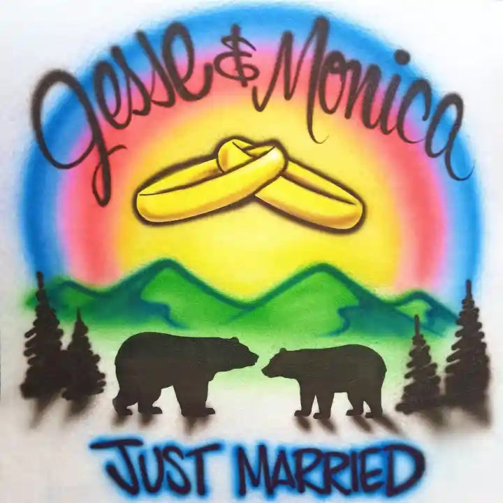 Airbrushed design featuring two crossed gold wedding rings under a rainbow gradient, with green mountains, black pine trees, two black bear silhouettes, and the text 'Jesse & Monica' and 'JUST MARRIED'.