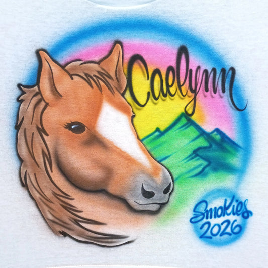 An airbrushed graphic on a white t-shirt. The design features a brown horse head with a white blaze, set against a colorful rainbow background with green mountains and the name 'Caelynn' in black script.
