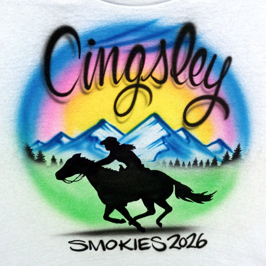 A white T-shirt with a colorful airbrushed graphic. The design features the word 'Cingsley' in large black script over a circular rainbow gradient background, with blue mountains, a black silhouette of a rider on a galloping horse, pine trees, and the text 'SMOKIES 2026' at the bottom.