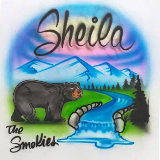 A design featuring a black bear standing beside a blue river with a small waterfall, green grass, dark trees, blue mountains, and a purple-pink sky. The word 'Sheila' appears at the top in large black script, and 'The Smokies' appears at the bottom left.