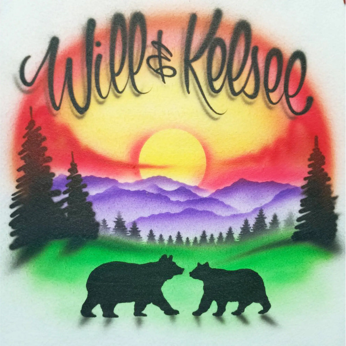 A colorful airbrushed-style graphic with the words 'Will & Kelsee' in black script over a sunset landscape. The design shows a yellow sun, red and orange sky, purple mountains, green grass, black pine trees, and two black bear silhouettes at the bottom.