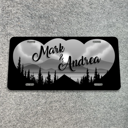 A black license plate with rounded corners, featuring a grayscale heart-shaped mountain and lake landscape with black tree silhouettes. The names 'Mark & Andrea' are printed in black script across the center.