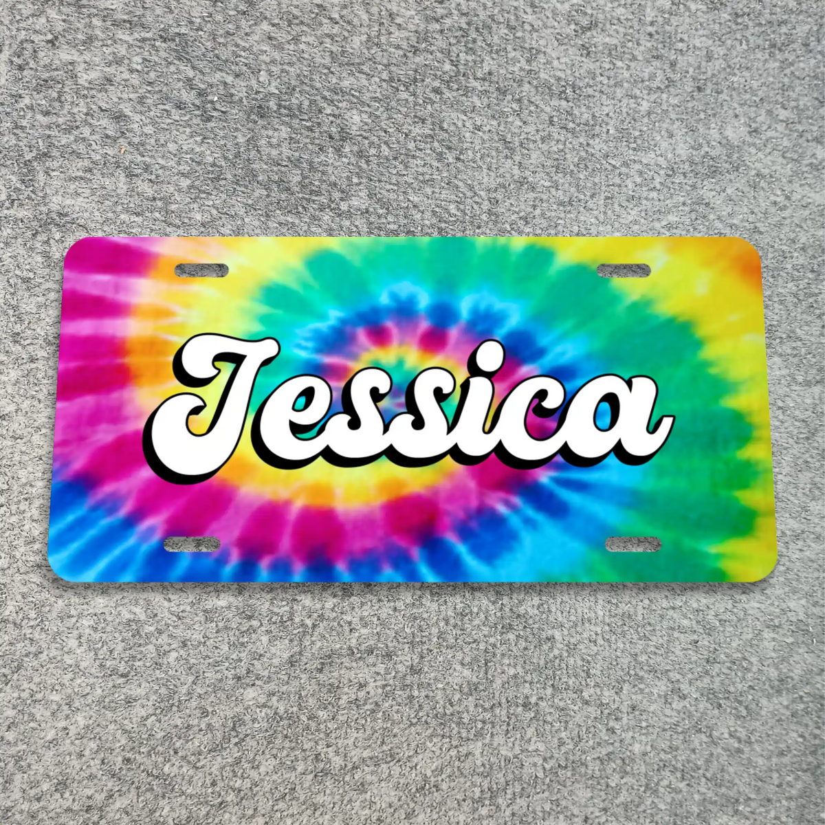 A license plate with a bright tie-dye spiral pattern in pink, blue, green, yellow, and purple, featuring the name 'Jessica' in large white cursive letters with a black outline across the center. The plate is photographed flat on a gray textured carpet background.