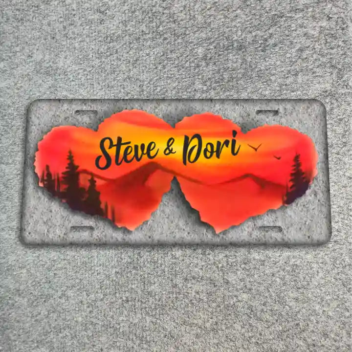A license plate featuring two overlapping heart-shaped cutouts filled with a sunset landscape design in orange, red, and yellow, with black silhouettes of trees and the text 'Steve & Dori' in black script. The plate is photographed flat on a textured gray surface.