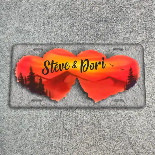 A license plate featuring two overlapping heart-shaped cutouts filled with a sunset landscape design in orange, red, and yellow, with black silhouettes of trees and the text 'Steve & Dori' in black script. The plate is photographed flat on a textured gray surface.