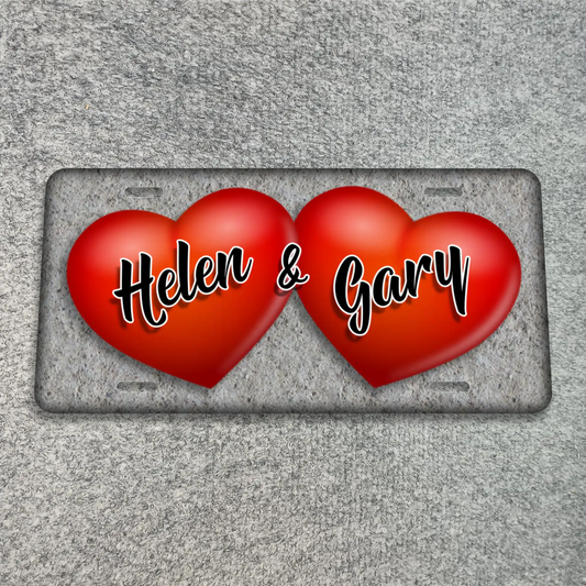 A textured gray license plate featuring two glossy red hearts side by side, with the names 'Helen & Gary' written across the hearts in white cursive with black outlines. The plate is shown on a textured gray background.