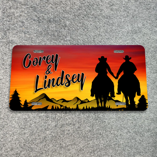 A license plate with rounded corners, featuring a sunset landscape design in red, orange, and yellow with black silhouettes of two cowboys on horseback, trees, and the text 'Corey & Lindsey'. The plate is photographed flat on a gray textured surface.