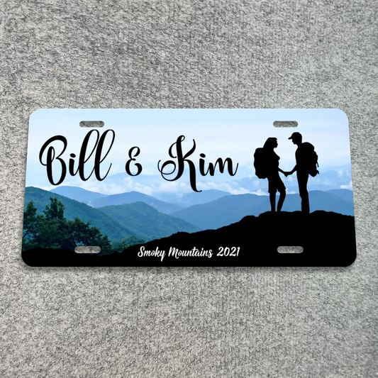 A custom novelty license plate featuring a scenic mountain landscape with the text 'Bill & Kim' and 'Smoky Mountains 2021', plus a black silhouette of a couple holding hands on a hill. The plate is photographed flat on a gray textured surface.