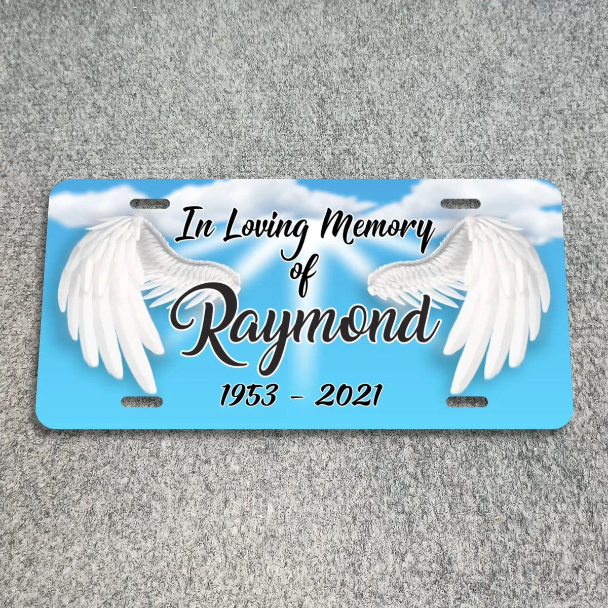 A personalized license plate memorial featuring a light blue sky background with white clouds and two large white angel wings. The text reads 'In Loving Memory of Raymond 1953 - 2021' in black script and serif lettering.