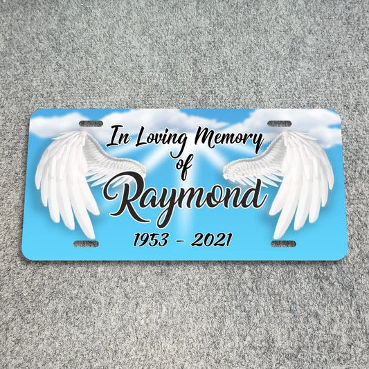A personalized license plate memorial featuring a light blue sky background with white clouds and two large white angel wings. The text reads 'In Loving Memory of Raymond 1953 - 2021' in black script and serif lettering.