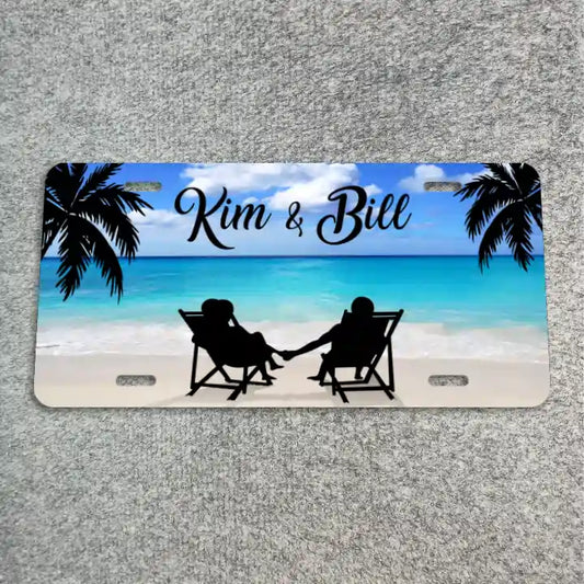 A license plate featuring a tropical beach scene with palm trees, turquoise water, white sand, and black silhouettes of a couple sitting in chairs holding hands. The text 'Kim & Bill' is printed across the top in black script.