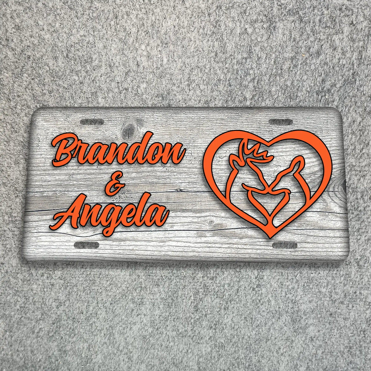 A personalized license plate with a weathered gray wood-grain background, featuring the names 'Brandon & Angela' in orange script and an orange heart outline containing a rose and two figures, displayed on a gray textured surface.