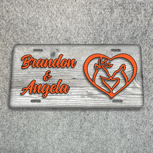 A personalized license plate with a weathered gray wood-grain background, featuring the names 'Brandon & Angela' in orange script and an orange heart outline containing a rose and two figures, displayed on a gray textured surface.