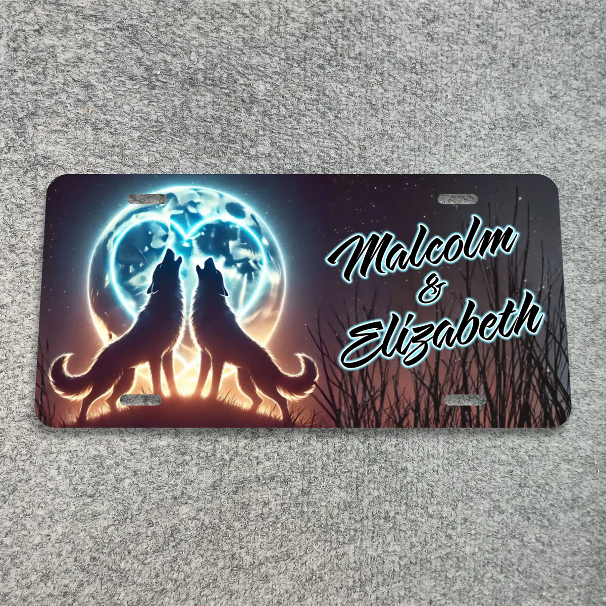 A custom license plate featuring a printed design of two howling wolves in front of a glowing full moon, with the names 'Malcolm & Elizabeth' in stylized white script. The plate is photographed flat on a textured gray fabric background.