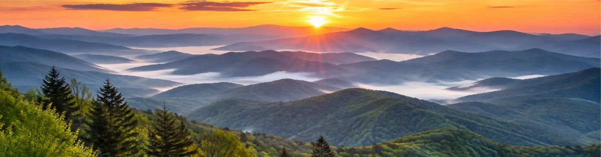 A scenic landscape print of a mountain range at sunrise, featuring layers of green forested hills, misty valleys, and a bright orange and yellow sun low on the horizon.