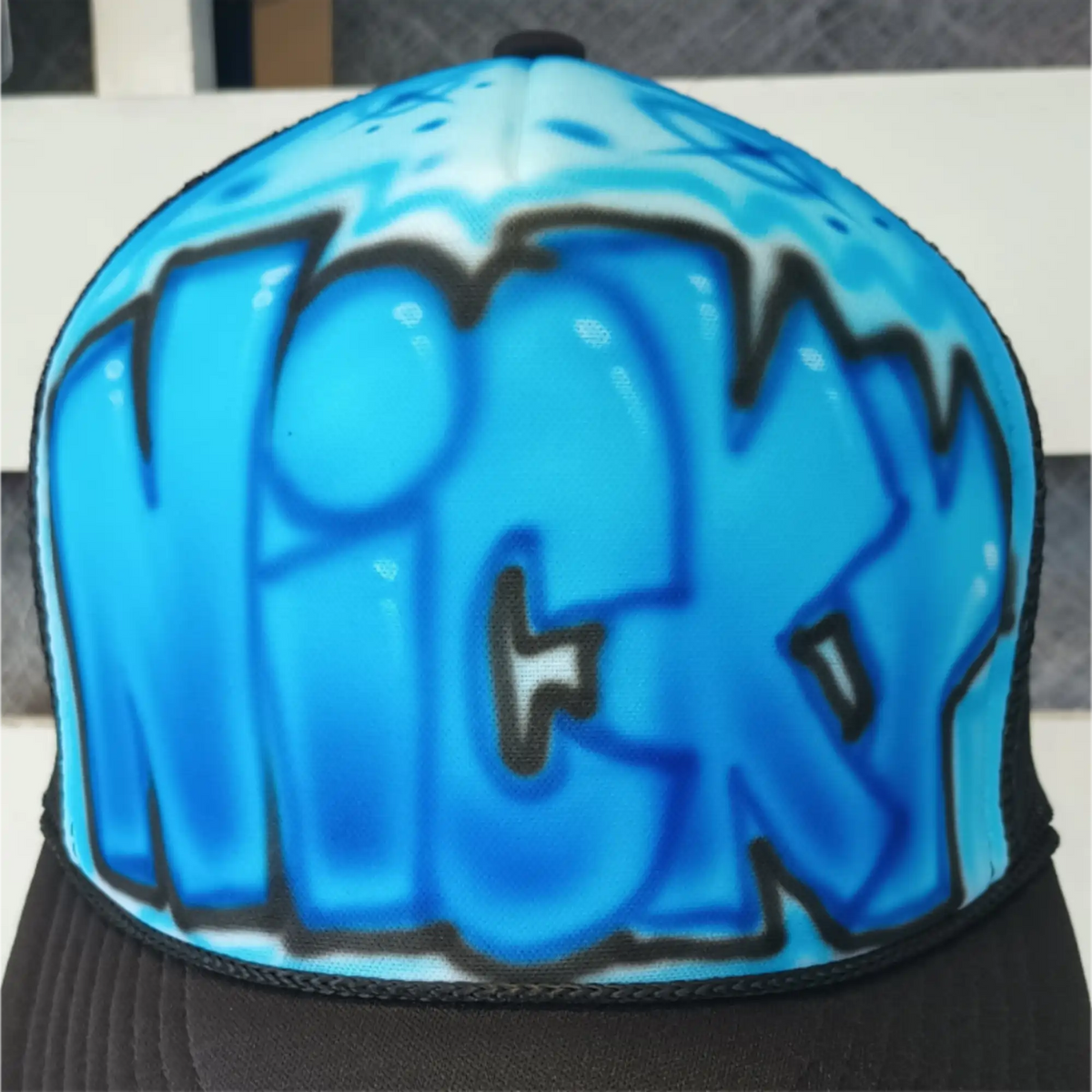 An airbrushed trucker hat featuring a bright blue graffiti-style 'NICKY' graphic with black outlines on the front panels, with a black 
brim.