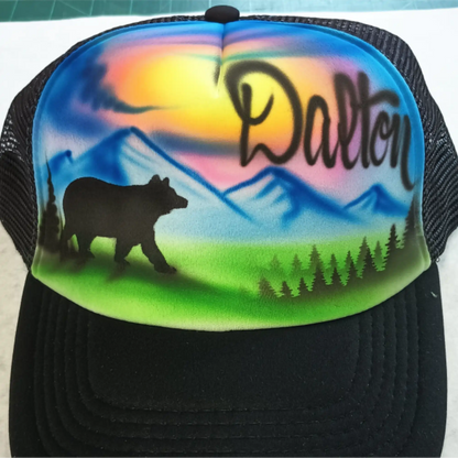 A black trucker hat with a colorful custom airbrushed front panel featuring a mountain sunset landscape, a bear silhouette, pine trees, and the name 'Dalton' in script.