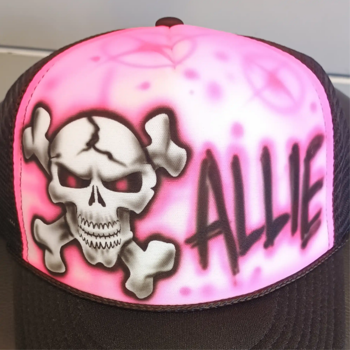 A trucker hat featuring a bright pink airbrushed front panel with a white skull and crossbones graphic, red eyes, and the name 'ALLIE' in black graffiti-style lettering. The brim and mesh side panels are black.