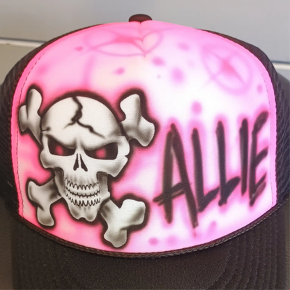 A trucker hat featuring a bright pink airbrushed front panel with a white skull and crossbones graphic, red eyes, and the name 'ALLIE' in black graffiti-style lettering. The brim and mesh side panels are black.